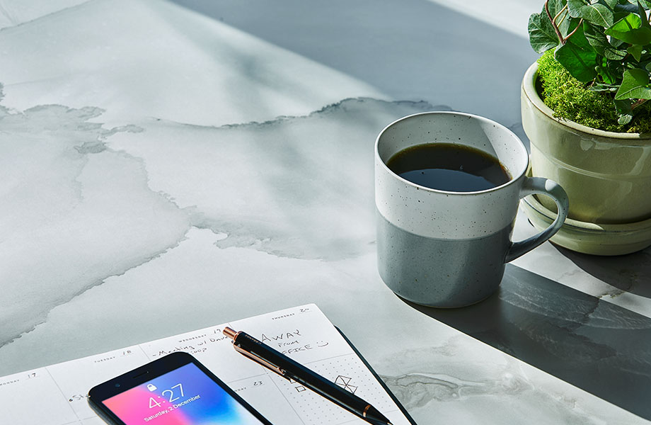 Table surface made with Formica® HPL laminate 180fx® 5016-11 Watercolor Porcelain - a soft blend of gray and beige, creating pockets of colour in the translucent overlaps. Cup of coffee, plant, phone and notebook