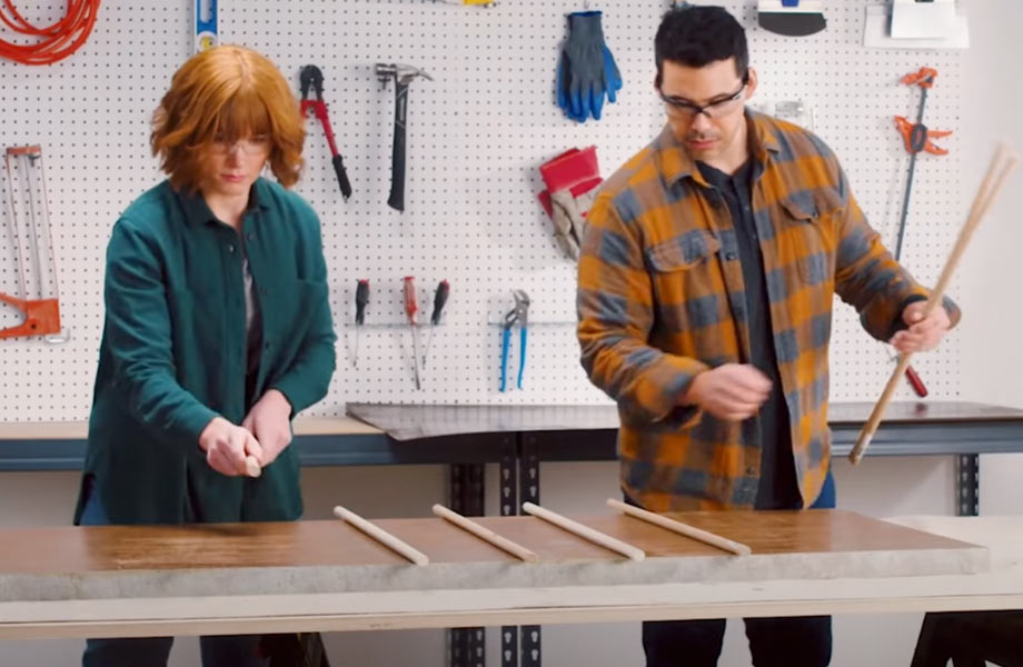 Man and woman in safety goggles lay dowel rods on work table in front of pegboard wall with tools