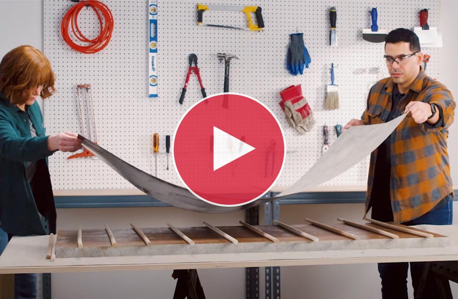 Man and woman in safety goggles lay sheet of Formica Laminate on work table in front of pegboard wall with tools 