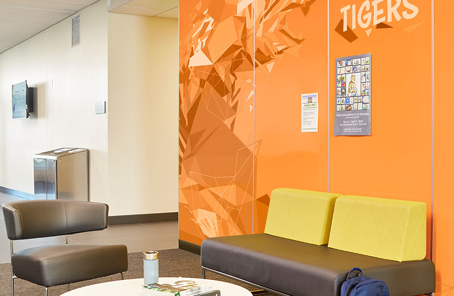 Aire d’attente avec canapé et chaise contre des panneaux Envision™ HardStop® de marque Formica de couleur orange qui mettent de l’avant un design dans le secteur de l’éducation et valorise une marque.