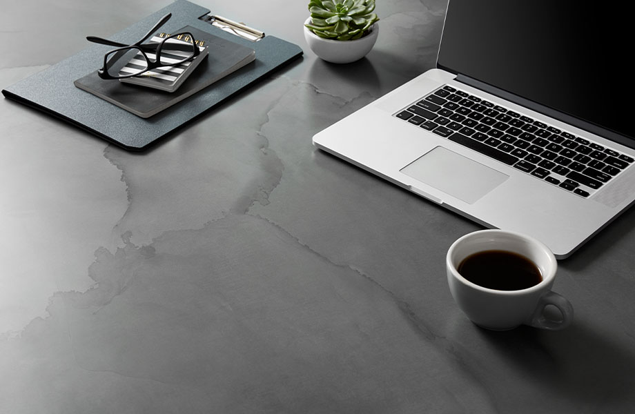 Table surface made with Formica® HPL laminate 180fx® 5017-11 Watercolor Steel – a blend of warm and cool grays with hints of blue and charcoal black as seen in tempered steel, a perfect pattern for the moody and inky tones in interiors today. Cup of coffee, notebook, plant, glasses, laptop